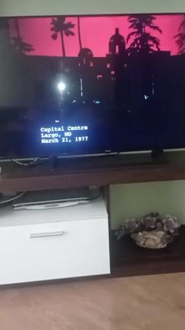 The video displays a television screen showing a bright, abstract, blue and white graphic. The graphic appears to be a stylized cloud or a smear of paint, with soft edges and varying levels of brightness. The screen is set against a dark background, likely a room, with a television stand below it. On the stand, a silver laptop is visible on the left, next to a white drawer unit. To the right of the laptop and below the screen, a decorative bowl filled with artificial flowers, in shades of pink and brown, sits on a darker surface. The lighting in the room appears dim, with the bright television screen being the primary light source. The floor beneath the stand is made of wooden planks. The time of day and weather are not discernible. No text is visible in the video. The overall scene suggests a quiet moment in a home environment.