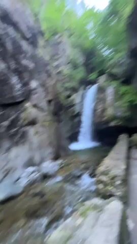 A person with dark hair in a braid, wearing a white outfit, walks toward a waterfall. The waterfall is surrounded by rocky cliffs and lush green trees. The person pauses to look up at the waterfall. The camera is held by the person, giving a first-person perspective. The video is blurry in some parts due to movement.