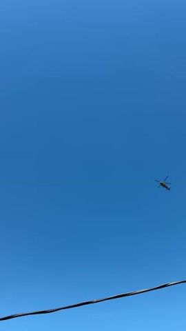 A military helicopter flies across a clear blue sky. The helicopter is a dark color, likely gray or black, and has rotating blades. A thin wire is visible in the foreground, possibly from a utility pole or fence. The helicopter appears to be moving from left to right in the frame.