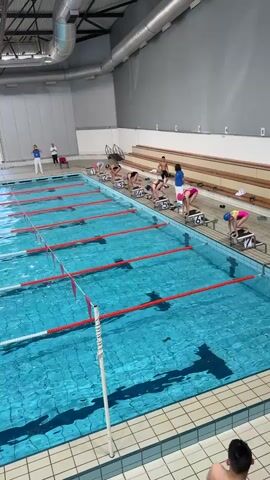 In an indoor swimming pool, eight swimmers are positioned on starting blocks, ready for a race. The pool is rectangular, with seven lanes marked by red-and-white lane ropes. The water is a clear, bright blue. The swimmers are wearing a mix of swimsuits and caps, and they appear to be young athletes. After a starting signal, they dive into the water simultaneously. Two adult figures dressed in athletic wear stand at the pool's edge, presumably coaches or officials. A second tier of bleachers sits to the side. The pool area has a high ceiling with industrial ductwork. Sunlight streams in through a large window at the far end of the pool.