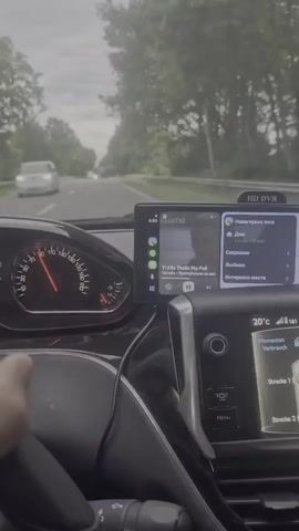 A first-person perspective video captured from inside a car shows the dashboard. The speedometer needle is just past the 80 mark. A navigation screen displays a map on the right side and music controls on the left. The song playing is "Ti Alle Thuis" by Fia Pii Pii Valadis. The car's climate control is visible below the navigation screen, showing a temperature of 20 degrees Celsius. Outside the car, a two-lane road is visible with trees lining both sides, and the sky is overcast.