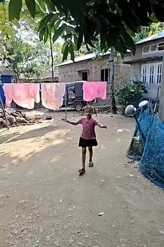 A young girl with pigtails, wearing a pink t-shirt and a black skirt, dances and walks in a dirt path between two buildings. The buildings appear to be made of corrugated metal and brick. Clothes are hanging on a clothesline in the background. A tricycle is parked near one of the buildings. The girl is smiling and appears to be enjoying herself. The scene is outdoors during the day.