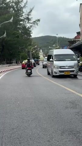 The video shows a first-person perspective of riding a scooter on a road in Karon, Thailand. The road is paved and has yellow lane markings. On the left side of the road, there is a sidewalk with red and white striped curb, followed by trees and a building in the distance. On the right side of the road, there are several parked motorcycles, businesses with awnings, and a multi-story building with a striped facade.

Several vehicles are visible on the road, including a white van, a silver car, a red car, and other scooters. A person on a scooter is in front of the camera. Pedestrians are seen walking on the sidewalk on the left side of the road.

The weather appears overcast, suggesting it might be late afternoon or an overcast day. The scene is bustling with traffic, indicating it's a public road in a populated area. The overall atmosphere seems to be a typical day in a tourist or commercial district.

No visible text can be deciphered from the video.