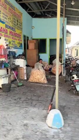 The video shows the exterior of a small workshop or repair shop. A person is sitting on the ground amidst large sacks of what appear to be snacks or food items. The workshop has a blue wall with a sign that says "BENGKEL USAHA MANDIRI". There are various objects scattered around, including tools, machinery, and other items associated with a workshop. Several motorcycles are parked outside the entrance. The ground is paved with interlocking bricks, and there is a covered area in front of the workshop. The lighting suggests it is daytime, possibly late afternoon.