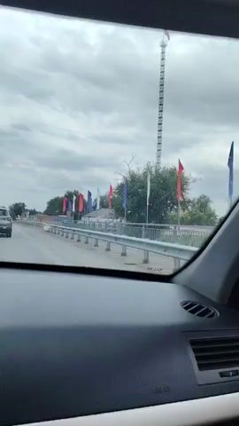 The video, recorded from inside a car, shows a person driving on a road with a cloudy sky overhead. A black car is visible ahead, and a cell tower can be seen in the distance. Flags of red, white, and blue are planted along the side of the road, and there are buildings with colorful geometric patterns. The driver's reflection in the rearview mirror shows them wearing sunglasses. The car's interior, including the dashboard and a side mirror, is also visible.