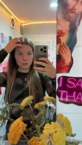 A young woman with long brown hair is taking a selfie in a mirror. She is wearing a dark, long-sleeved shirt and a necklace. The mirror is in a room decorated with a string of lights, stickers, and a poster of Jesus. There is a vase of yellow flowers in front of the mirror, and reed diffusers on a surface below. The woman is holding her phone in her right hand and looking at the screen. The background wall is covered in doodles and stickers.