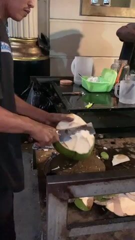 A man wearing a black short-sleeved shirt with a blue and white pattern on the left sleeve and a watch on his left wrist is shown from the waist up. He is standing in front of a metal table with a coconut resting on it, which he is opening with a large knife. The coconut is mostly green with a white top and some brown peel remaining on the sides. There are other coconut parts on the table. To the right, another man is partially visible. Behind the table, a white pitcher and a green bowl are visible. The lighting is dim, suggesting it is either evening or night.