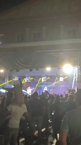 A video captures an outdoor concert at night. A stage is set up with a banner that reads "ТРАДИЦИОНЕН ЛУКОВИЧКИ ПАНАИР" in Cyrillic, indicating a traditional Lukovichi fair. Two performers are on stage, one in a white shirt and baseball cap rapping or singing, and another in a black dress playing a saxophone. Disco balls hang above the stage, reflecting bright yellow and white lights that illuminate the performers and the audience. The crowd is densely packed, with many people raising their hands in the air. In the foreground, a person is being carried on someone's shoulders, their back to the camera, and they appear to be holding a small flag. The atmosphere is lively and festive.