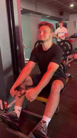 A young man with light brown hair and a black t-shirt and shorts is sitting on a weight machine in a gym, pulling a cable towards himself. He is smiling. In the background, another man with a shaved head and a teal t-shirt and shorts walks past. The gym has red lighting and weight plates are visible on racks.