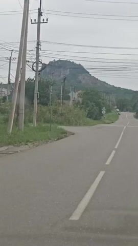 The video captures a view from a moving vehicle on a paved road. The road curves to the left, leading towards a large, rugged mountain in the distance. The mountain has a rocky, uneven surface with sparse vegetation. Power lines crisscross the sky above the road and extend towards the mountain. On the left side of the road, there are utility poles and dense green foliage. On the right side, the road continues uphill, with more trees and some structures visible further up. The sky is overcast, suggesting a cloudy or hazy day. There are traffic markings on the road, including a dashed white line separating lanes and a solid white line on the edge of the road. A few traffic signs are also visible along the roadside. The overall scene depicts a rural or semi-rural landscape with a prominent natural landmark.