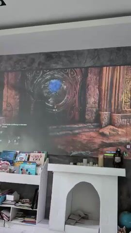 The camera is positioned in a room where someone is playing a video game on a large screen projected onto the wall. The game features dark, stone architecture and a character exploring a dimly lit area. Below the screen, there's a white bookshelf filled with books and other items, and next to it, a small white decorative fireplace. A lantern sits atop the fireplace. The bookshelf contains books like "SUPER ZOA" and "ATLAS," among others. The room is well-lit, with a white ceiling and what appears to be a gray textured wall behind the projected screen. The focus is on the screen displaying the video game.