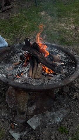 The video shows a metal fire pit with a fire burning inside. The fire pit is filled with ash and burning wood. A person in jeans places a small log upright in the fire pit. The background consists of grass and dirt. The fire is burning with small flames and smoke.