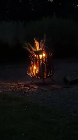 The video shows a bonfire burning in a metal fire pit at dusk or night. The fire is contained within a decorative, openwork metal structure supported by three legs. The flames are bright orange and yellow, casting a warm glow on the surrounding area. Smoke rises from the fire, dissipating into the dark air. The ground around the fire pit is covered in dark gravel and some patches of grass. In the background, a dark line of trees or bushes is visible against the night sky. The scene suggests an outdoor setting, possibly a backyard, campsite, or park, given the presence of the fire pit and natural elements. The time of day is clearly nighttime or late evening due to the darkness. There is no visible text in the video. The overall mood of the video is calm and serene, evoking the cozy atmosphere often associated with campfires.