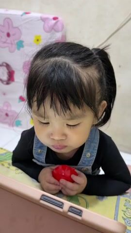 A young child with dark hair pulled back in a ponytail is sitting at a table, looking down at a pink tablet. They are wearing a black long-sleeved shirt and blue denim overalls. In their hands, they are holding a small red object. To the right of the child, a person's hand is holding a spoon and a clear plastic container filled with food. The person is spoon-feeding the child, who opens their mouth to receive the food. After eating, the child closes their mouth and looks away from the spoon. The background shows a patterned surface, possibly a bedspread or tablecloth.