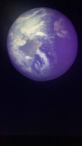 The video shows a circular object against a black background that appears to be changing from a blue-purple rendition of the Earth to a purple moon with craters. The moon's appearance is rough and heavily textured with many small indentations. The scene is dimly lit.