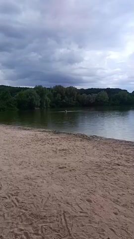 A sandy beach with people and a lake. In the foreground, there is a sandy beach with footprints. In the middle ground, there is a body of water with some people swimming and a few individuals sitting on the shore. In the background, there are trees and a cloudy sky. The overall scene depicts a recreational area by a lake on a cloudy day.