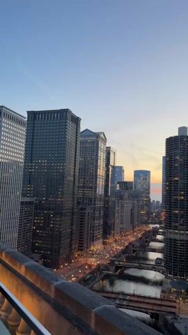 The video displays an evening cityscape in Chicago with numerous skyscrapers, a river, and bridges. The sky transitions from light blue to orange at the horizon. The lights from the cars and buildings illuminate the scene. The Trump International Hotel and Tower is visible with its name displayed.