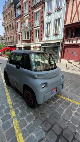 A grey Citroen Ami electric vehicle is parked on a cobblestone street. The car has a black roof and black trim around the windows. The wheels have red accents. The street has yellow painted lines. In the background, there are buildings with traditional architecture, featuring colorful flags strung across. A sign on one of the buildings reads "GALERIE FONTAINE". The scene appears to be in a European town or city.