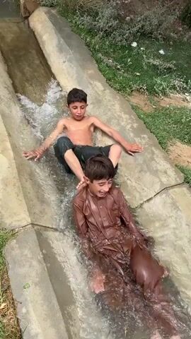The video shows a group of boys and an adult sliding down a concrete water channel. The channel is open at the top and appears to be filled with flowing water, creating a makeshift water slide. The environment surrounding the channel is dry and dusty, with sparse vegetation. The people are dressed in casual clothing and are clearly enjoying themselves, splashing and sliding in the water. The camera angle is from above, looking down at the scene, and the video is shot in daylight.
