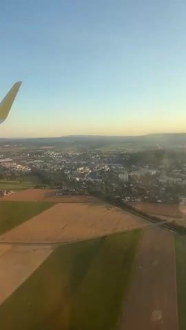 The video shows an aerial view from an airplane window. The left side of the frame is dominated by the wing of the airplane, which is white with a yellow tip. Below the wing, a patchwork of agricultural fields is visible, with some fields appearing golden brown and others green. In the distance, a town or city is spread out, with buildings and roads. The sky is clear and blue, with a gradient towards a lighter hue near the horizon. The lighting suggests it is either sunrise or sunset, casting a warm glow over the landscape. The camera is stationary, offering a static perspective of the scene. There are no people visible in the frame.