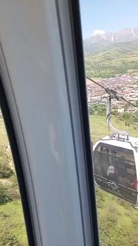 The video shows a view from inside a cable car as it descends towards a station. The cable car in view is white with a dark window, and appears to have people inside. Below, there is a green, grassy hillside with some trees, and a building with a dark gray roof. Beyond that, there is a river, and a town with many houses and buildings. In the distance, there are mountains with snow on top.