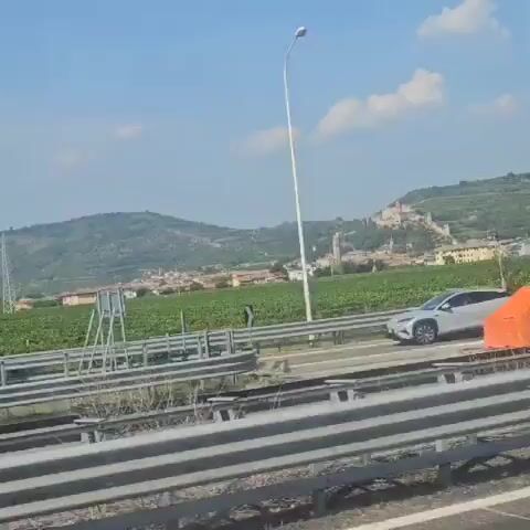 The video pans across a landscape with a highway in the foreground, bordered by guardrails. An orange traffic cone is visible on the side of the road. Beyond the highway, a green field, possibly vineyards, stretches out. In the distance, a village with buildings and a tall clock tower is nestled on a hillside. Dominating the hilltop above the village is a large, ancient-looking castle complex with stone walls and towers. The sky is blue with scattered clouds. The overall impression is of a scenic rural view with historical architecture.