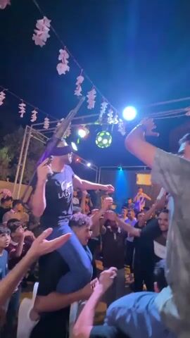 The video shows a nighttime outdoor gathering with a group of people, some of whom are dancing. Several individuals are riding on the shoulders of others. One person, sitting on someone's shoulders, is holding up a rifle. Colorful lights, including red, blue, and green, illuminate the scene. There are decorations hanging overhead, resembling paper lanterns. In the background, a band or musicians can be seen performing on a stage. The overall atmosphere appears to be celebratory.