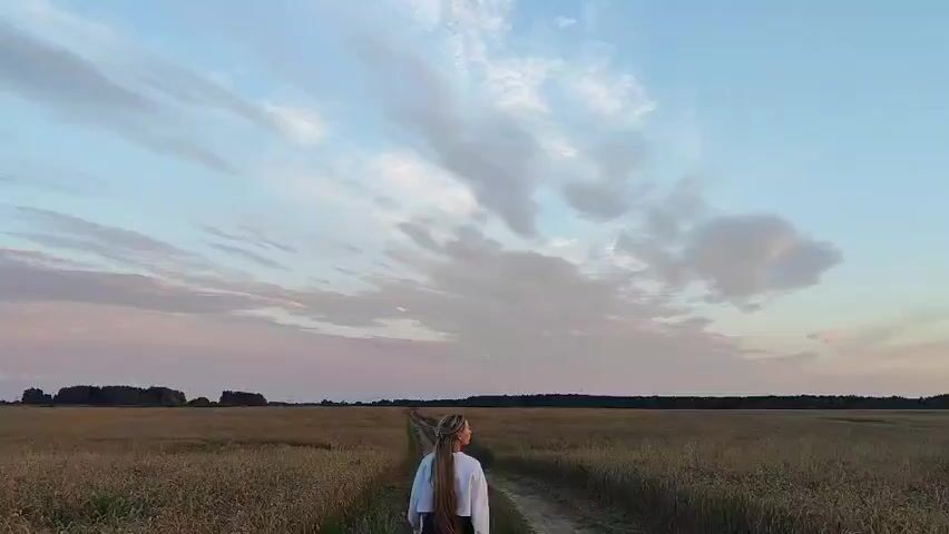 A person with long blonde hair, wearing a white hoodie, stands facing away from the camera on a dirt path that cuts through a field of wheat. The field extends to the horizon, where it meets a line of trees. The sky above is a pale blue, with wispy clouds tinged with pink and orange from a setting sun. The person is positioned in the center of the frame, on the path, with the field on either side and the expansive sky above. The overall scene is one of peaceful solitude in a rural landscape during twilight.