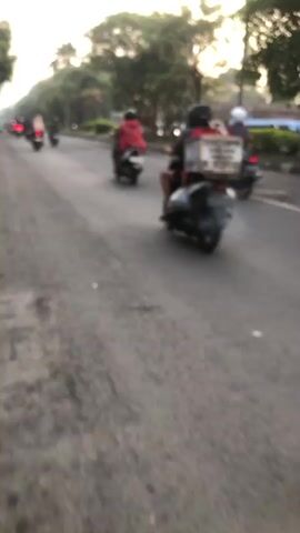 The video shows a first-person perspective of someone riding a scooter or motorcycle on a paved road. The rider is wearing a light-colored helmet and an orange t-shirt. The scooter is off-white with red accents. In front of the rider, several other motorcycles and scooters are visible, also carrying riders wearing helmets. The road appears to be lined with trees and some buildings or structures are present in the background. The lighting suggests it is either early morning or late afternoon. The overall scene depicts a common mode of transportation in an urban or suburban environment.