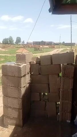 A video shows a collection of concrete blocks stacked to create a makeshift doorway or structure. In the background, there is a rural landscape with some buildings under construction, piles of dirt or sand, and trees. The sky is visible, with some clouds. A person's legs and feet, wearing sandals and patterned socks, are visible on the right side of the frame, suggesting someone is standing or sitting there. The camera is positioned at a low angle, looking upwards towards the sky and the constructed blocks.