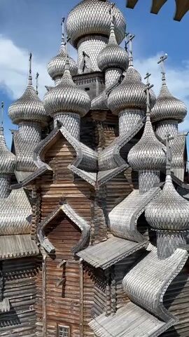 The video shows a close-up view of a wooden church with multiple onion domes, a traditional architectural style. The camera pans to the left, revealing a scenic landscape with a large body of water, lush green trees, and a pathway where several people are walking. The sky is blue with fluffy white clouds, indicating fair weather. The church itself is made of logs, and its intricate wooden carvings and tiered domes are clearly visible. The overall impression is one of historical architecture set against a natural, serene backdrop.