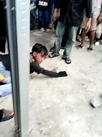 A person is lying on the ground near a metal pole, surrounded by onlookers. The person on the ground is wearing a black t-shirt and light-colored pants. They appear to be reaching out. A black cloth object is on the ground nearby. Several people are standing and observing the scene. Some are wearing black clothing. The ground is a rough, concrete-like surface. In another segment, people are gathered around a figure, some of whom are wearing black outfits.