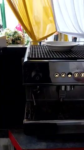 Here is a detailed description of the video:

The video shows a person making coffee using an espresso machine. The espresso machine is stainless steel with several buttons and levers. The person is using the machine to dispense coffee into a stainless steel cup. The counter space around the machine is cluttered with coffee beans, milk, and other items. There are yellow and white curtains in the background.