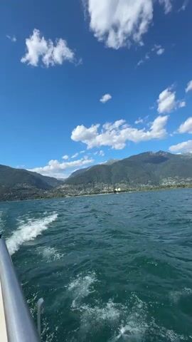 This video captures a scenic view from a boat moving on a body of water, likely a lake, given the mountainous landscape in the background. The water is a vibrant turquoise, with white frothy wakes trailing behind the boat on the left side of the frame, indicating forward motion. The sky is a brilliant blue, dotted with fluffy white clouds.

In the distance, a picturesque town or city is nestled along the shoreline, with buildings climbing up the gentle slopes of the surrounding mountains. The mountains themselves are covered in lush green vegetation, and some appear to have snow-capped peaks further in the distance. The overall impression is one of a clear, sunny day, likely in the late morning or early afternoon, given the brightness of the light and the shadows.

The setting is a beautiful natural landscape, with the calm waters of the lake contrasting with the imposing mountains. The activity is simply enjoying a boat ride and taking in the scenery. There are no people or specific objects visible besides the boat railing on the left edge of the frame and the distant buildings. The mood conveyed is peaceful and serene, evoking a sense of vacation and relaxation. There is no visible text in the video. The location is identified as Locarno, Switzerland, which is known for its beautiful lake and surrounding mountains.