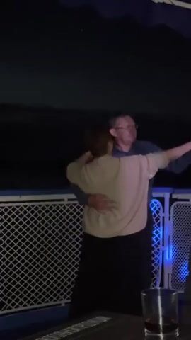 A man and a woman are dancing together at night. The woman is wearing a tan sweater and a black skirt, while the man is wearing a blue shirt and dark pants. They are holding each other and appear to be in a romantic embrace. The background is dark and appears to be outdoors, possibly on a boat or a deck, with a railing and some lighting visible. There is a glass with a dark liquid on a surface in the foreground.
