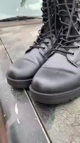 Two black combat boots with laces rest on a surface, presumably a vehicle hood given the windshield's edge in the background. The boots have a leather-like material on the lower portion, with fabric uppers laced to the top. The surface they rest on appears to be painted dark green, with some flaking or weathering evident. The overall setting suggests an outdoor environment, likely on a cloudy day given the muted lighting.