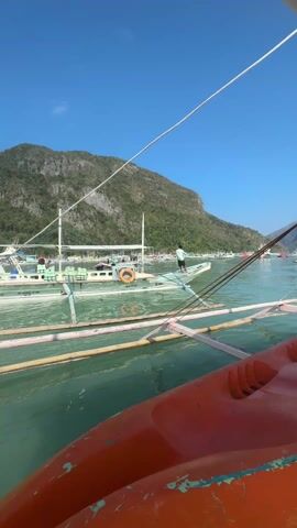Here's a concise description of the scene:

The video shows a view from a boat on calm, light green water. Several outrigger boats are visible, some with people on them. In the foreground, there is a bright orange kayak secured on the deck of the boat from which the video is being taken. A person wearing a pink life vest is seated, visible in the lower right corner.  In the background, a lush green mountain rises, and the sky is clear blue.