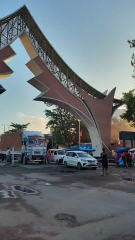 The video shows a large, modern archway with a metallic grid structure, possibly at a border crossing or entrance to a complex. Several large trucks, including a flatbed and a tanker truck, are parked in front of the arch. Smaller white vehicles, possibly cars or vans, are also present. A few people are visible, some standing and observing, others appearing to be involved in activities related to the vehicles. The ground is paved but has puddles of water, suggesting recent rain or damp conditions. The sky is clear and blue, indicating daylight. The overall scene appears to be one of a transportation hub or a point of entry with ongoing activity.