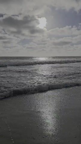 A video displays a body of water at the shoreline, with waves approaching a sandy beach. The sun is visible through clouds, casting a reflection on the water's surface. The overall tone of the video is muted and appears to be a still shot.