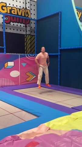 The video shows a man in a pink shirt and white pants jumping on a trampoline in an indoor park called Gravity Jump. He attempts a backflip but lands on his back. The surrounding area is decorated with colorful padding and geometric designs. A voice counts down "5 seconds" just before he attempts the flip.