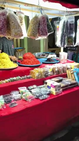 The video opens with a close-up of a market stall piled high with colorful candies and dried goods. The camera then pans across the stall, revealing more packaged sweets and snacks. A person, partially obscured, appears to be tending to the stall. The scene shifts to a busy street with heavy traffic. Cars and motorcycles are bumper-to-bumper, suggesting a traffic jam. Several people are visible, some on motorcycles and others standing by the roadside. The sky is overcast, indicating dusk or an evening setting. The overall impression is that of a lively, bustling marketplace with a lot of activity.