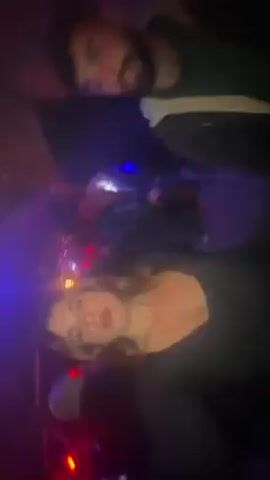 A woman with blonde hair dances in a crowded, dimly lit club. The lighting is colorful, with red, blue, and green lights flashing. Other people are seen dancing around her. She wears a dark, low-cut top and moves energetically to the music. The camera angle is low and tilted, giving a distorted view of the scene. A man with a beard appears briefly in the background.