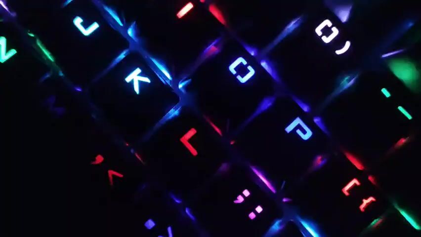 This is a close-up shot of a black keyboard with colorful LED backlighting. The keys are illuminated in various colors including blue, green, red, purple, and white, creating a vibrant and dynamic effect against the dark background. Some of the visible keys include letters such as 'M', 'K', 'L', and 'P', as well as punctuation marks. The lighting shifts and changes, highlighting different keys and sections of the keyboard. The focus is sharp on the keys, with a slight bokeh effect in the background, emphasizing the illuminated keyboard as the central subject.