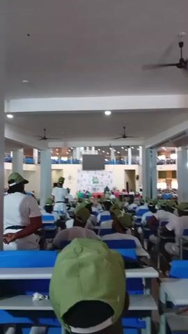 The video shows a large group of young people, likely young adults, seated at desks in a lecture hall or examination room. They are all wearing matching green caps and white shirts, suggesting they are part of an organized group or program. Many are looking forward, some appear to be listening to someone speaking at the front of the room. A few individuals in uniform, possibly instructors or invigilators, are visible walking among the seated attendees. The room is well-lit, with ceiling fans visible, and appears to be a modern, enclosed space. A large screen or projector is mounted at the front of the room. The overall atmosphere appears to be one of a formal gathering or educational event.
