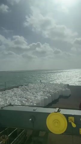 The video captures an elevated view of a bustling dock area on a sunny, partly cloudy day. In the foreground, a gray metal structure, possibly part of a crane or ship, features a yellow wheel and the inscription "SWL : 10/40 KN," indicating its Safe Working Load. Numerous large white bulk bags (FIBCs) are neatly stacked on the paved dock, stretching along the waterfront. Beyond the dock, a vast expanse of choppy, blue-green ocean reflects bright sunlight, creating a shimmering effect. As the camera pans slightly to the right, more of the dock is revealed, showing additional red plastic barriers or containers and a tall light pole. The sky is dotted with white clouds, with patches of bright blue visible. No people are visible, and no active loading or unloading is taking place, but the arrangement of the bags and equipment suggests ongoing or impending cargo operations in a port or harbor setting. The location is unknown, but the provided context states "Unknown, Colombia."