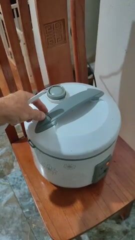 A person opens the lid of a rice cooker, revealing freshly cooked white rice inside. The rice cooker is white with gray accents and floral designs around the bottom. It sits on a wooden chair with an ornate back. The background includes a white wall and part of a porch railing. The person uses their left hand to lift the lid, which has a gray handle.