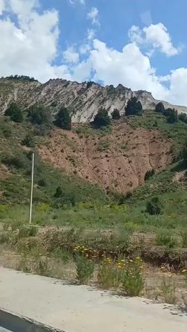 A video shows a mountainous landscape with green vegetation and sparse trees on the slopes. The sky is partly cloudy with blue sky visible. A white pole stands vertically on the hillside. A road or path is visible in the foreground. The overall impression is of a natural, undeveloped area.