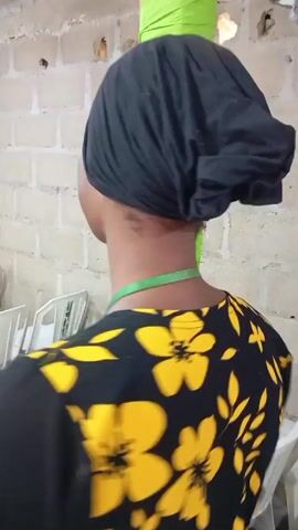 A woman with a black headwrap and a black shirt with yellow floral patterns walks through an indoor space. She has a green ribbon or lanyard around her neck. To her right, there is a stack of brightly colored fabrics (green and orange). In the background, rows of white chairs can be seen. Other people are also present in the space. The sound of music is audible. The room appears to be a large hall or meeting room.