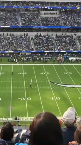 The video depicts a football game in progress at a stadium filled with spectators. The focus is on the field, where a kicker prepares to kick the ball. The field is marked with yard lines and the Chargers logo. The camera angle is from the stands, offering a wide view of the field and the surrounding crowd. People are seated in the foreground, observing the game.