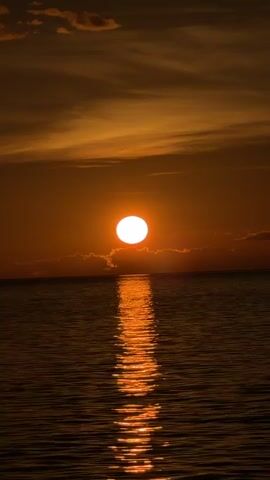 The video shows a sunset over the ocean. The sun is a bright white orb partially obscured by clouds at the horizon. A reflection of the sun creates a shimmering path across the dark water. The sky is filled with orange and brown clouds. The camera is still and does not move throughout the clip.